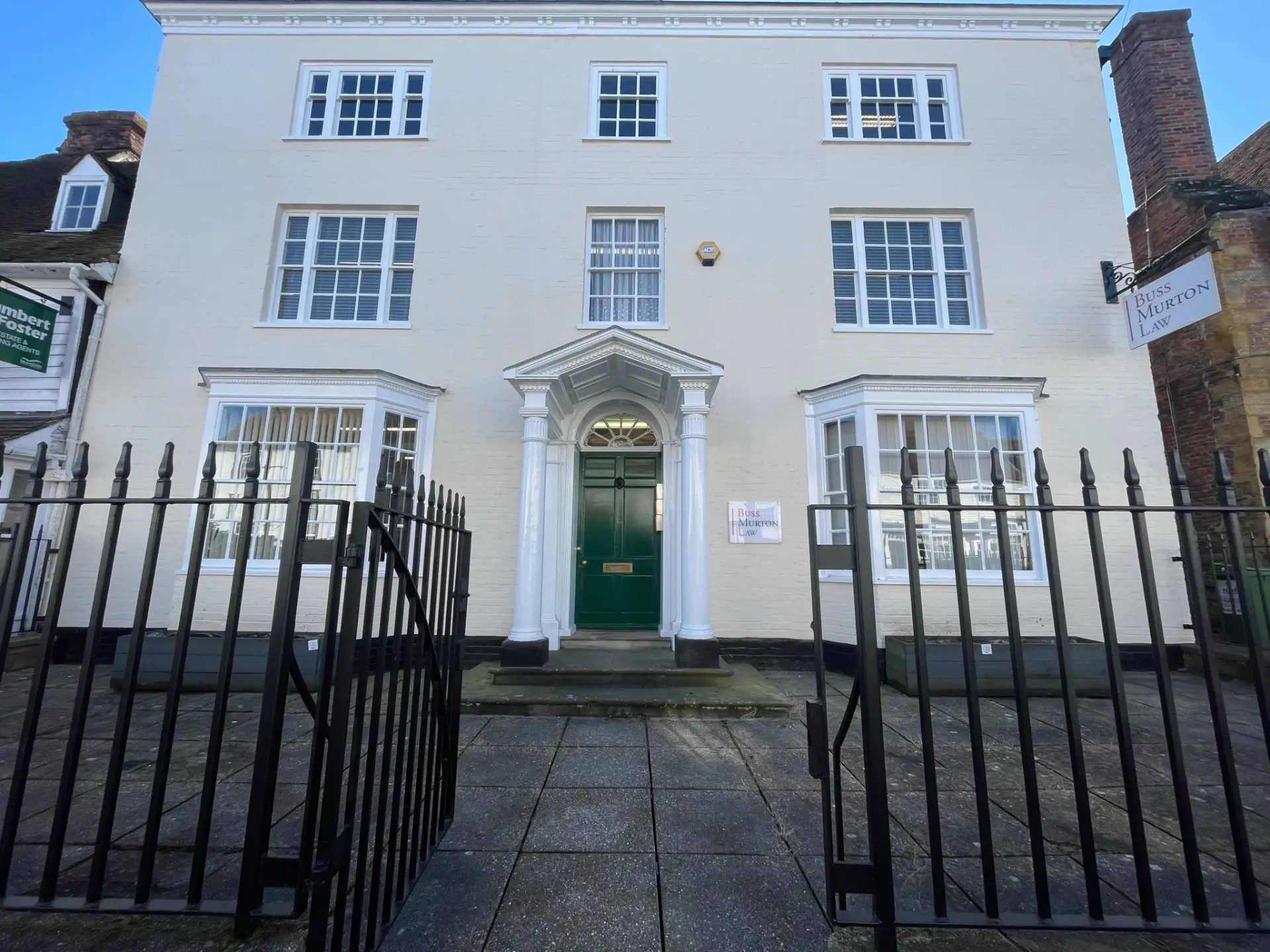 Clermont House on the High Street, Cranbrook: a Georgian three-storey cream-stuccoed townhouse, with a pillared classical doorcase, green front door, four-light sash windows in symmetrical pairs, and black wrought-iron railings at the pavement carrying a green Buss Murton plaque.