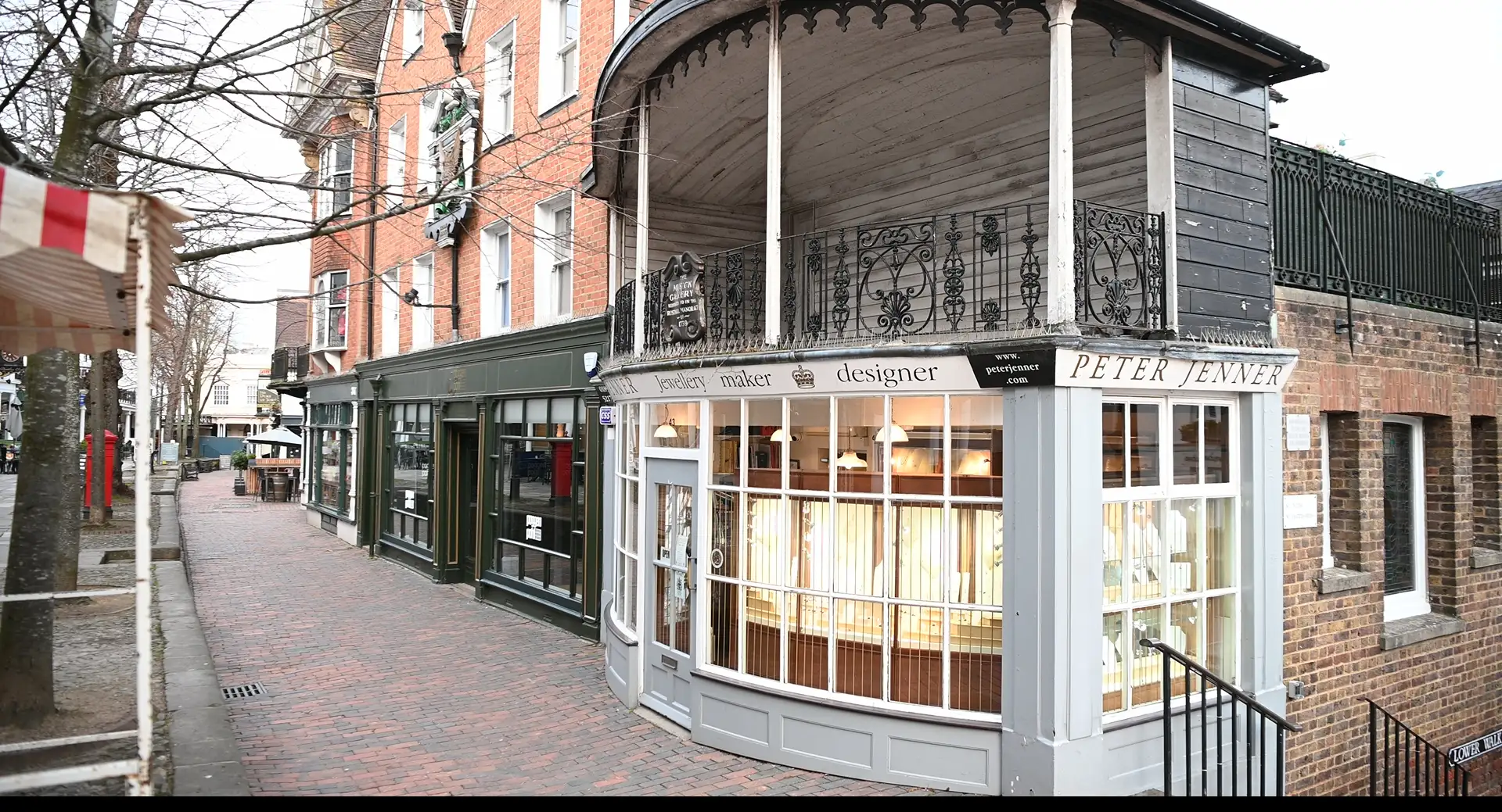 The Pantiles colonnade in Royal Tunbridge Wells: red brick Georgian shopfronts with white pilasters, a delicate wrought-iron Regency balcony above the central shopfront, and the worn-paved pedestrian street running along its length.